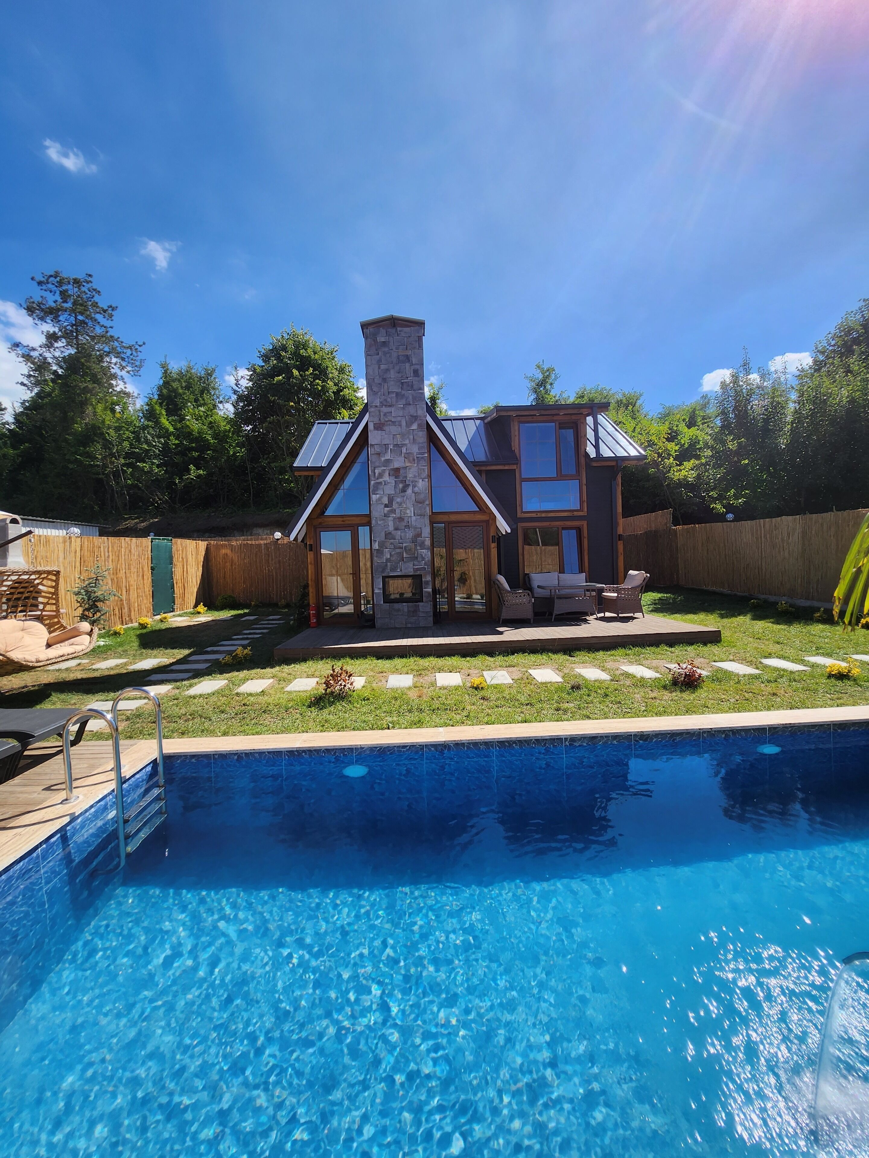 A modern house with a stone chimney, large windows, and a swimming pool in the foreground.