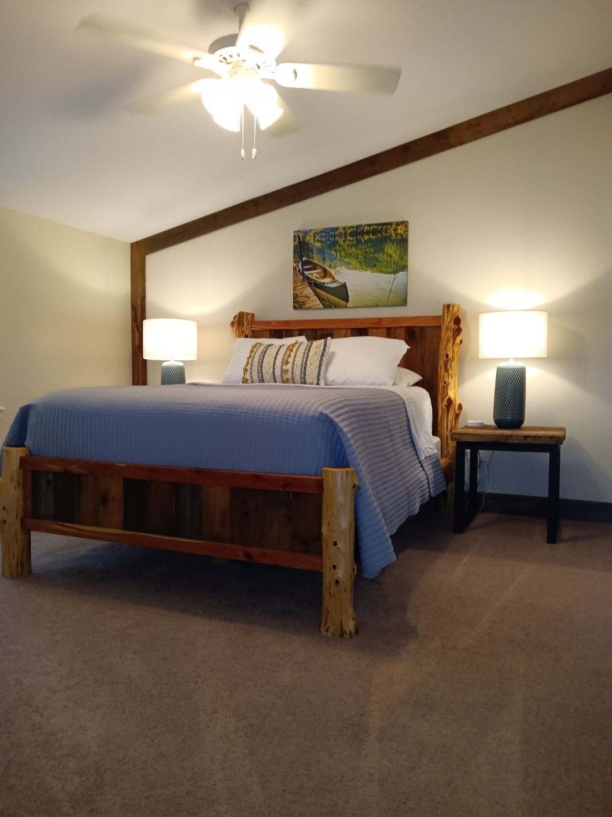 A bedroom with a wooden bed, two bedside tables with lamps, a ceiling fan, and a painting on the wall.