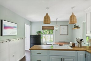 A kitchen with light blue cabinets, a wooden countertop, hanging wicker pendant lights, a window with a view, and a framed picture on the wall.