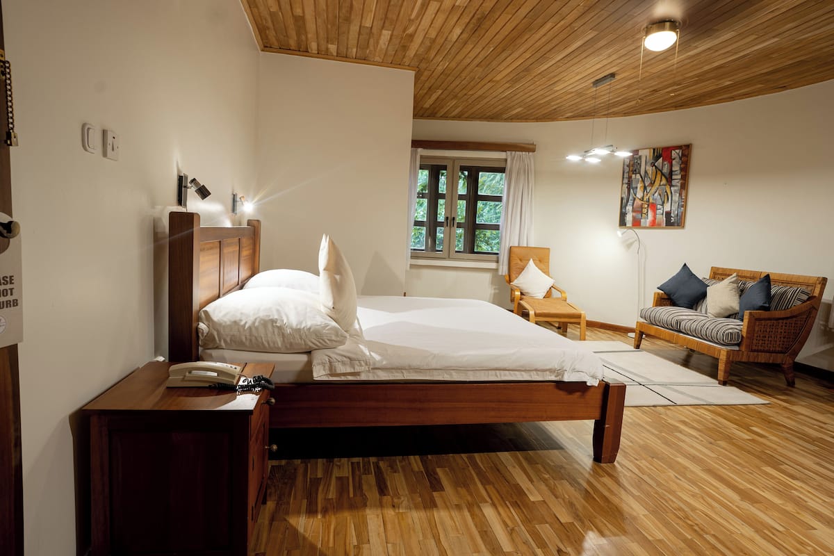 Un dormitorio con una cama de madera, una mesita de noche con un teléfono, una ventana con cortinas y un sillón de mimbre con cojines.