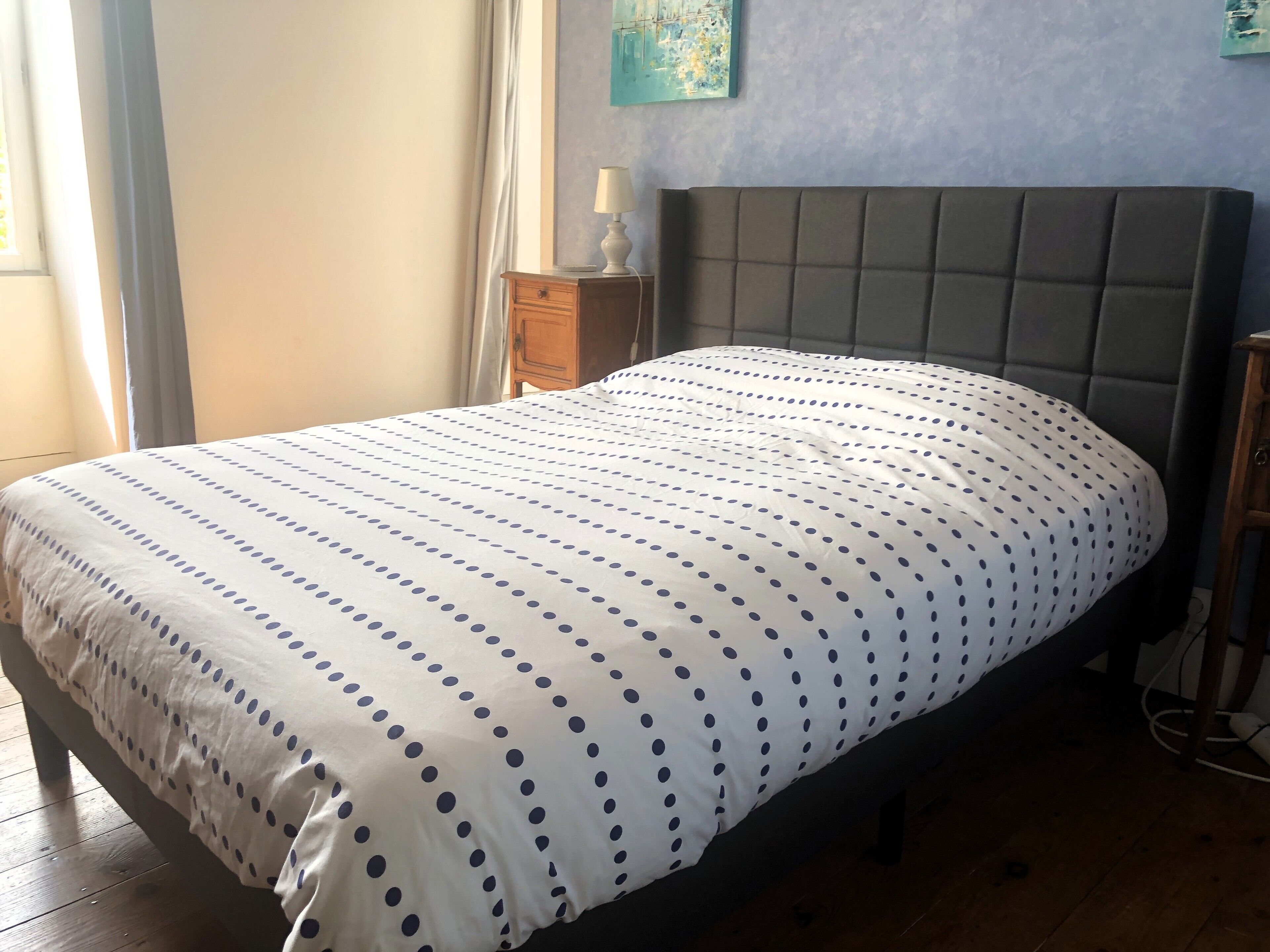 Una cama con una colcha de lunares blancos y azules, una mesita de noche de madera con una lámpara, y una pared azul con un cuadro.