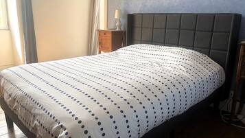 Una cama con una colcha de lunares blancos y azules, una mesita de noche de madera con una lámpara, y una pared azul con un cuadro.