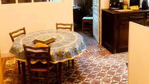 Un comedor con una mesa redonda cubierta por un mantel floral, rodeada de sillas de madera. El suelo presenta un diseño de baldosas con motivos.