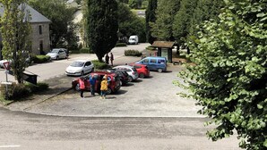 Un estacionamiento con varios coches y un autobús, rodeado de follaje y un edificio de fachada de piedra.