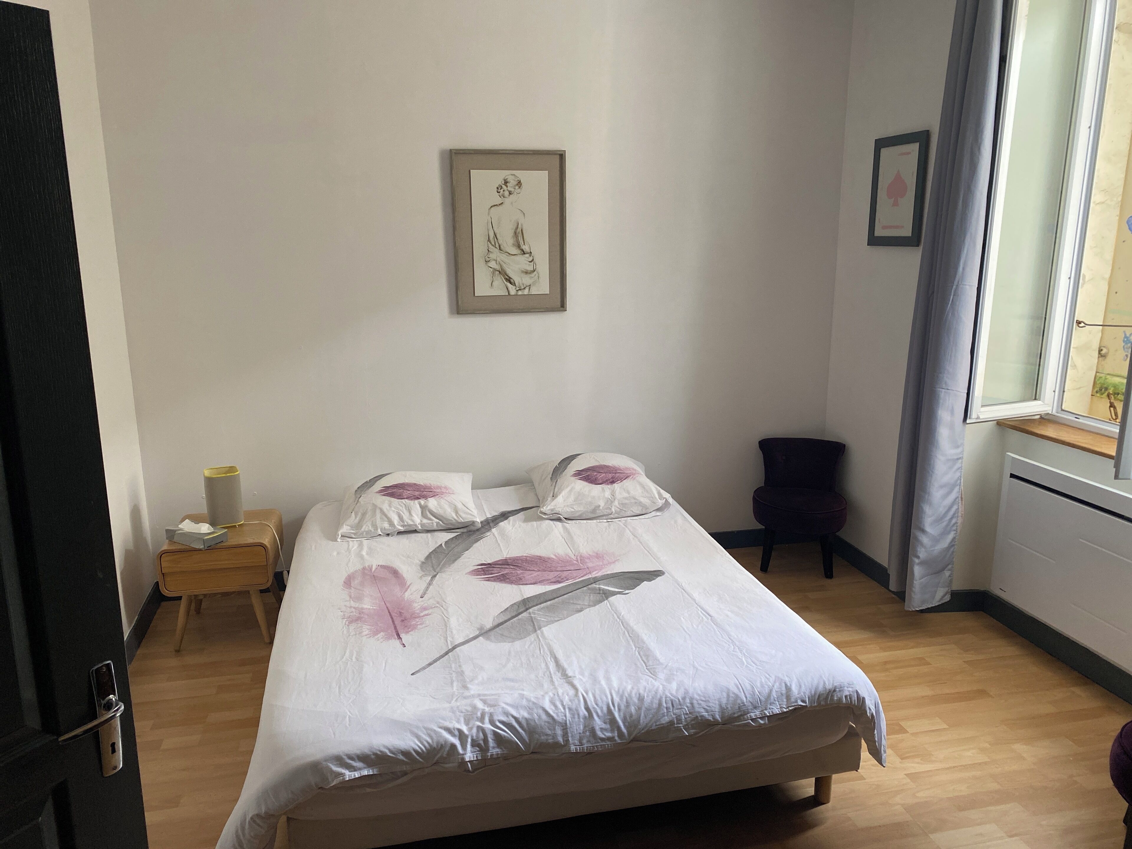 A bedroom with a bed, a nightstand, a chair, a window with curtains, and a framed picture on the wall.