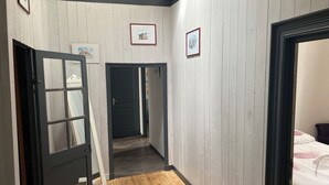 Ein Flur mit Holzboden, einer schwarzen Tür und einem Bett mit weißer Bettdecke im angrenzenden Zimmer.
