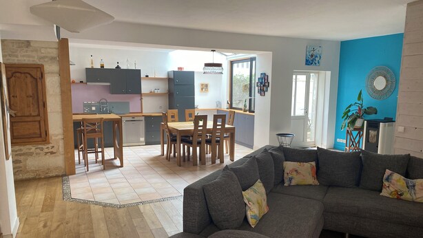 Ein modernes Wohnzimmer mit einem grauen Sofa, einem Holzesstisch und Stühlen. Im Hintergrund ist eine Küche mit grauen Schränken und einem Fenster zu sehen.