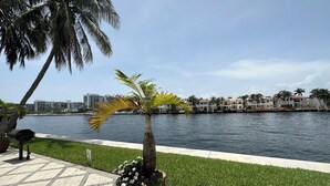 Une promenade en bord de mer bordée de palmiers, avec une zone herbeuse et une vue sur les bâtiments situés de l’autre côté de l’eau.