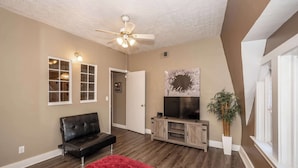 A living room with a black leather sofa, a TV on a wooden stand, a ceiling fan, and a potted plant.