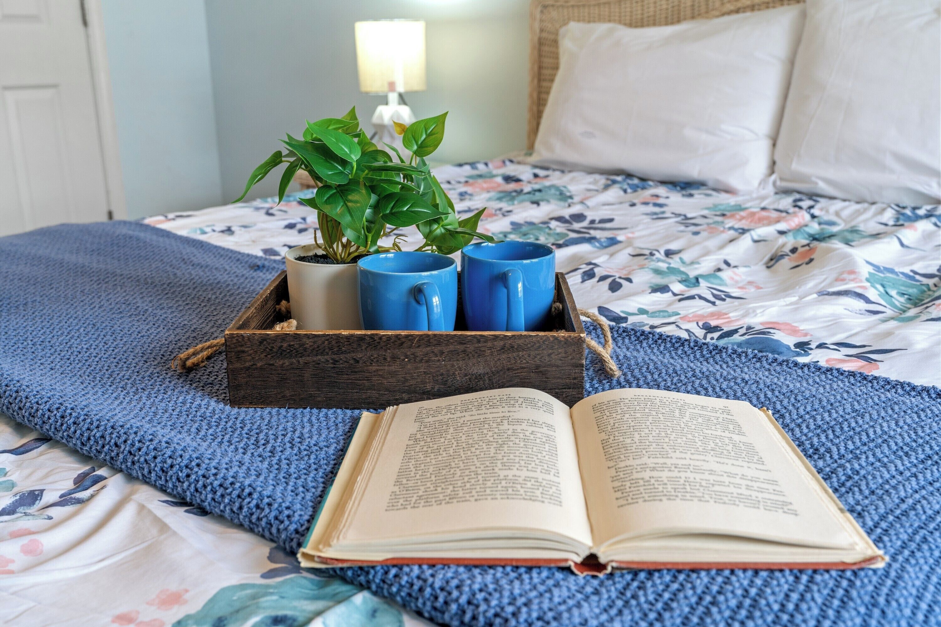 Una cama bien hecha con una colcha estampada, una manta azul de punto y una bandeja de madera con una planta en maceta y dos tazas azules.