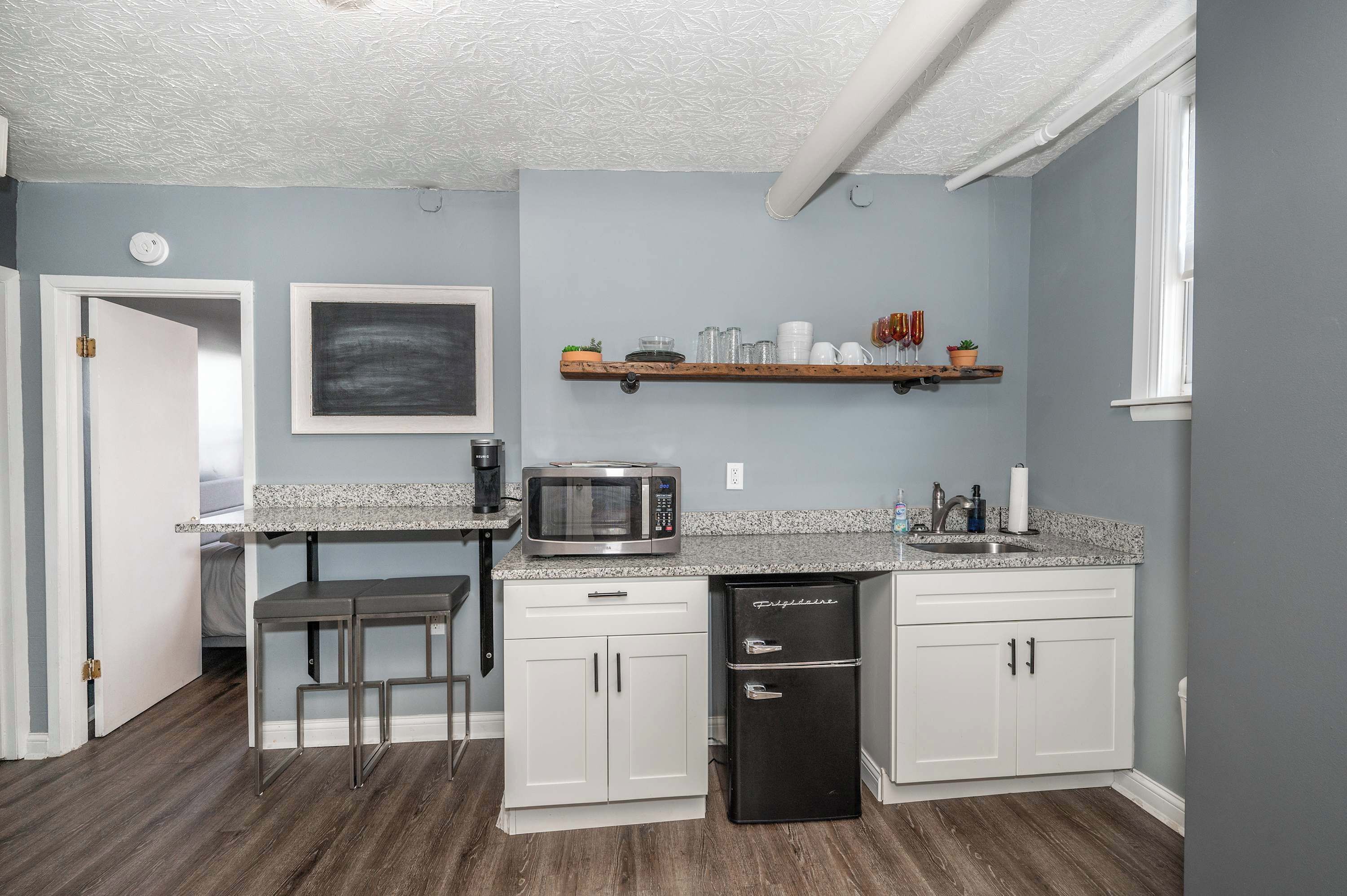 A compact kitchen with white cabinets, a granite countertop, a microwave, a refrigerator, and a small bar stool.