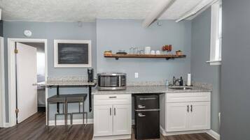 A compact kitchen with white cabinets, a granite countertop, a microwave, a refrigerator, and a small bar stool.