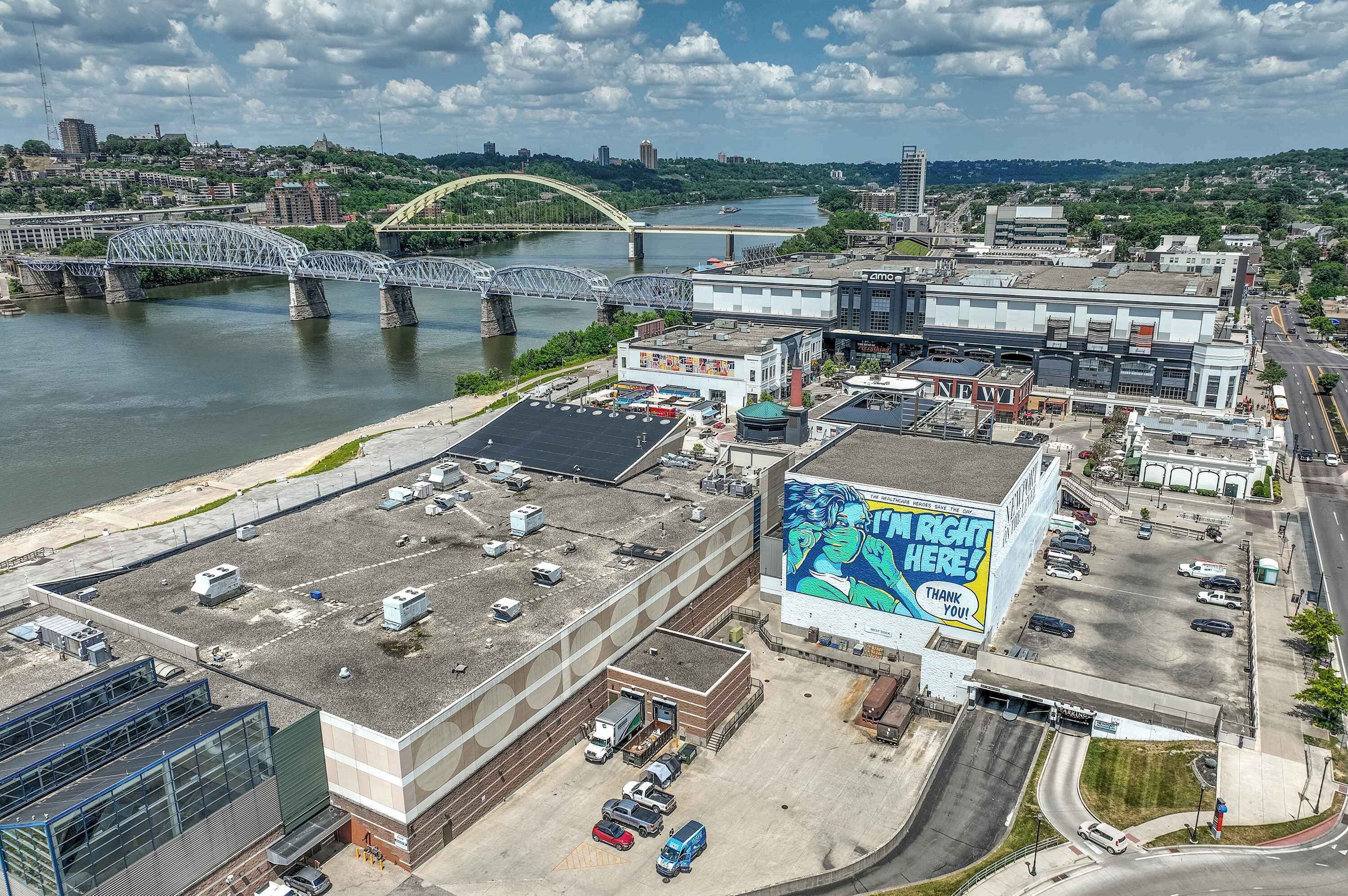 A commercial area with a large building featuring a prominent mural, a parking lot, and a bridge over a river.