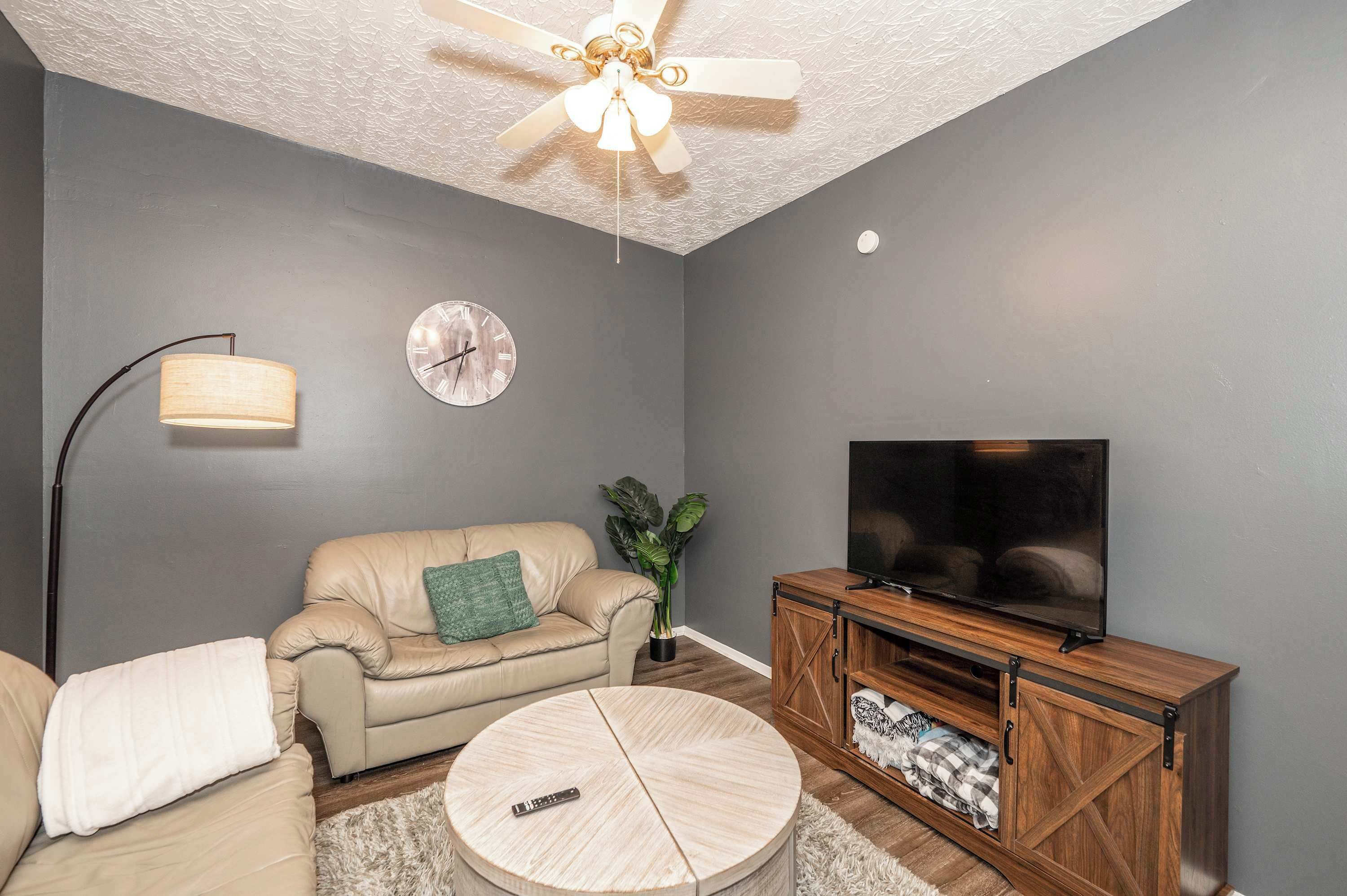 A living room with a ceiling fan, a clock, a television, a sofa, a coffee table, and a lamp.