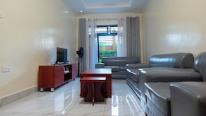 A living room with a sofa, a coffee table, a TV on a wooden stand, and a view of greenery outside. - VENUS APARTMENT KIGALI (Kigali)