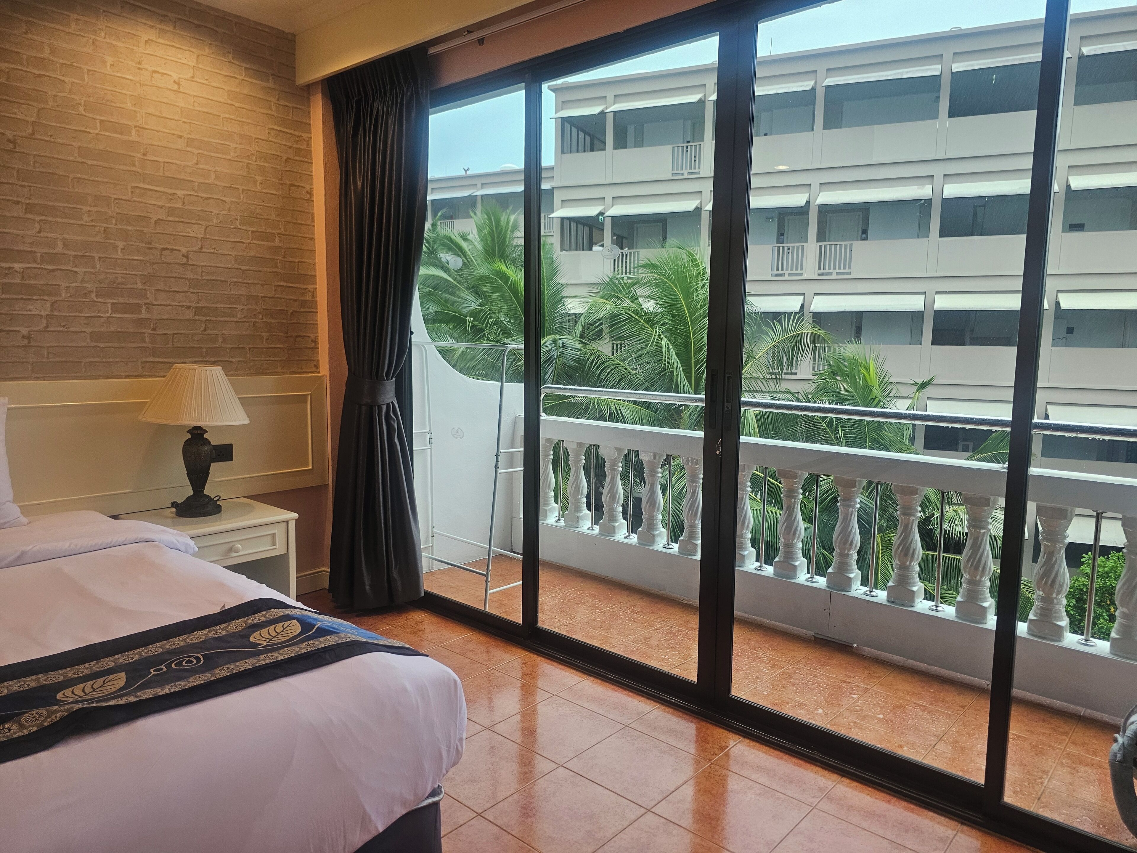 Una habitación de hotel con cama, mesita de noche, lámpara y ventana que ofrece vistas a un edificio y palmeras.