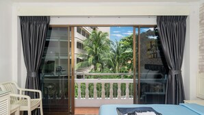 Habitación de hotel con balcón, cama, una silla y vistas a palmeras y edificios.