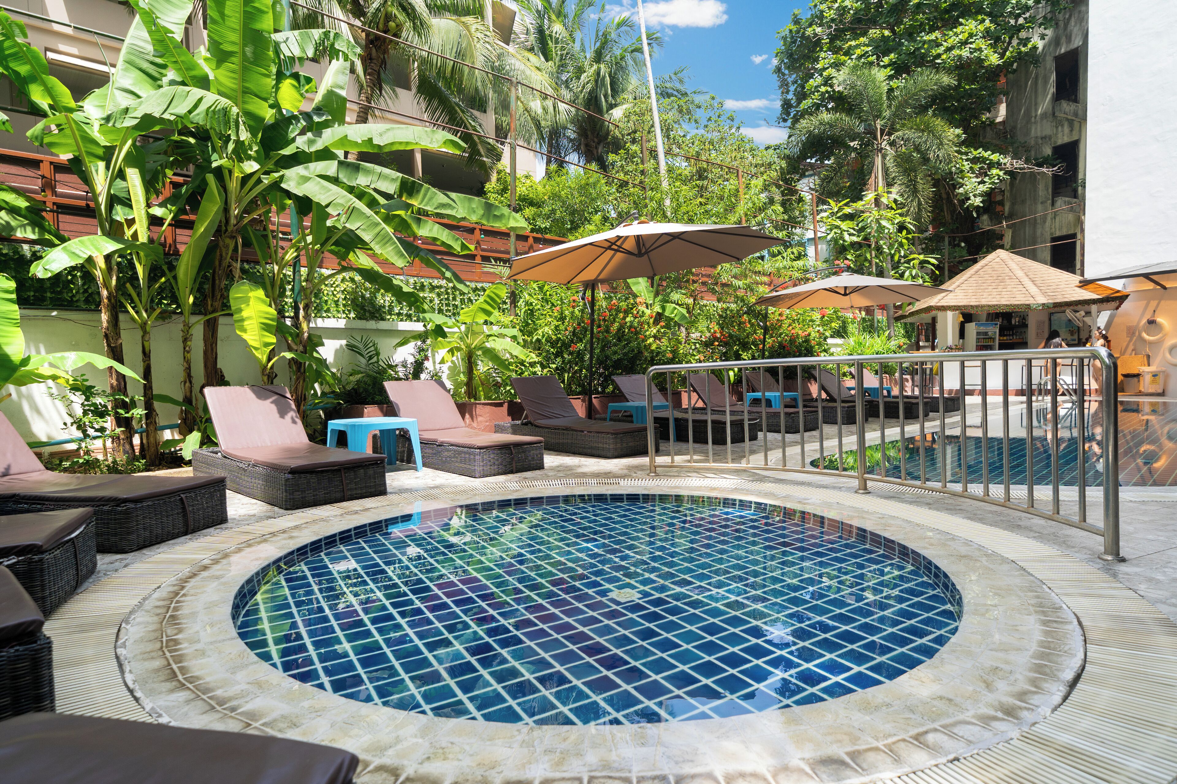 A circular pool with a tiled edge, surrounded by lounge chairs and umbrellas, with lush greenery and a building in the background.