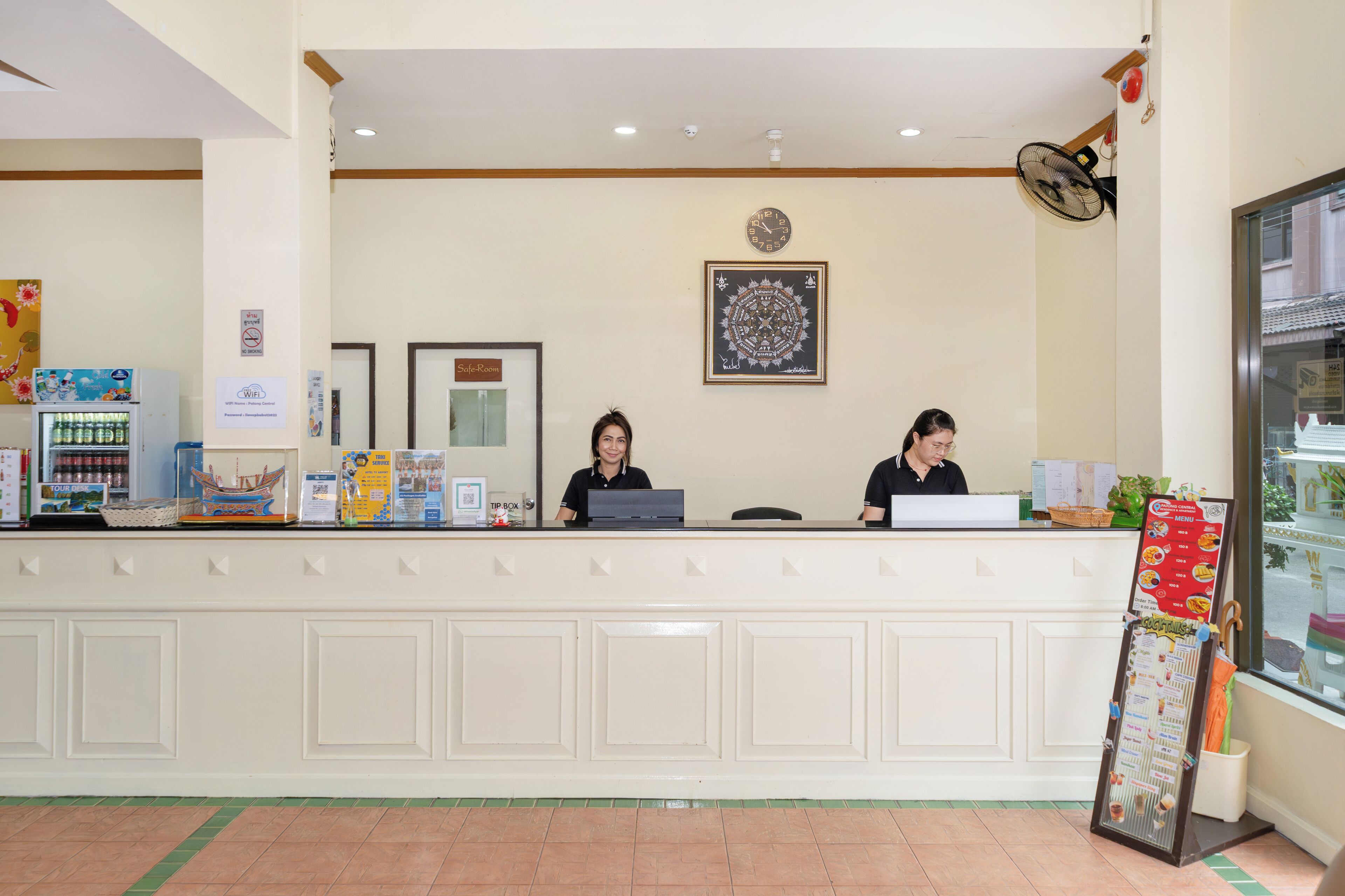 A reception desk with two staff members, a clock, a framed artwork, and a menu board.