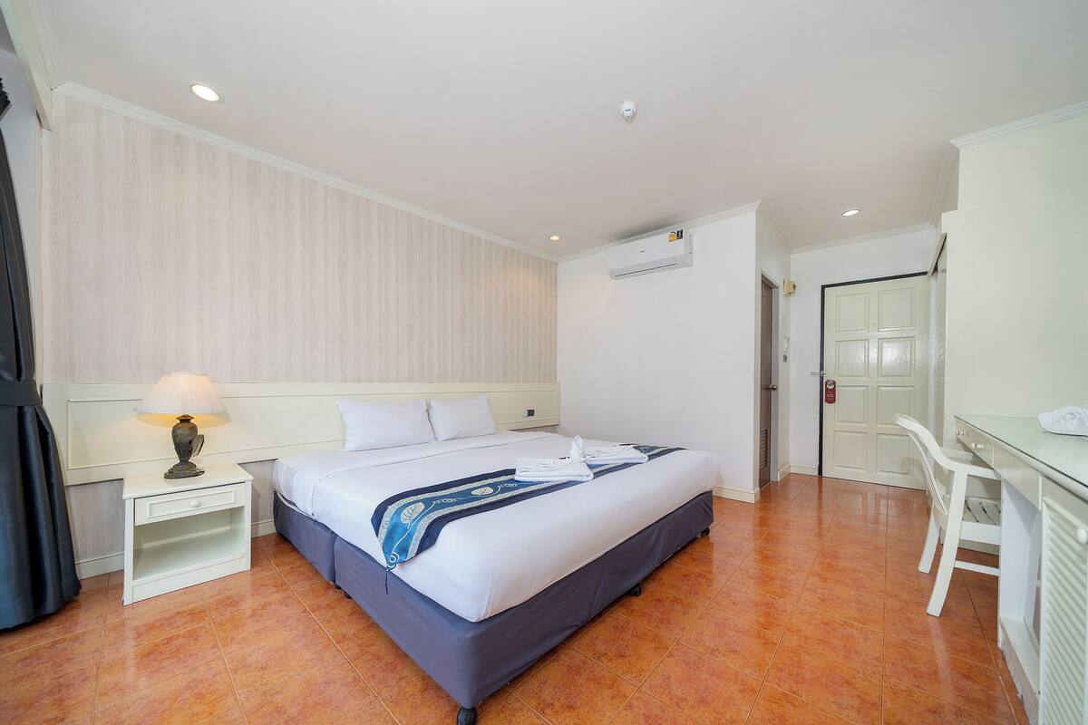 A hotel room with a bed, a desk, a chair, a nightstand, and an air conditioning unit.