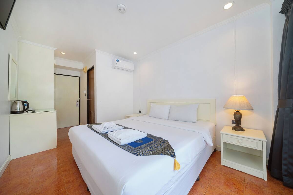 A hotel room with a large bed, a bedside table with a lamp, a small desk with a kettle, and an air conditioning unit on the wall.