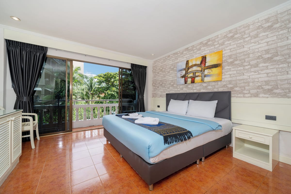 A bedroom with a large bed, a balcony with curtains, a white nightstand, and a brick-patterned wall.