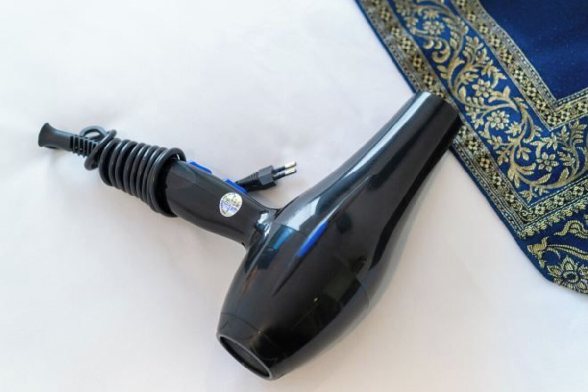 Un secador de pelo negro con enchufe y cable, sobre una superficie blanca junto a un tejido azul estampado.