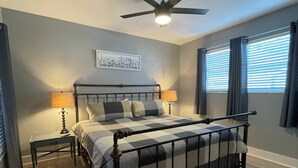 A bedroom with a bed, two bedside tables, a ceiling fan, a window with curtains, and a wall-mounted picture. - Melbourne, FL Home-Away From Home - Eau Gallie District  (Melbourne)