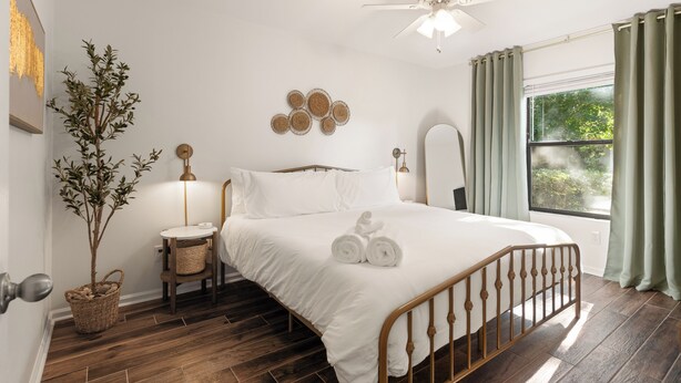 A bedroom with a wooden bed, a bedside table, a wall-mounted mirror, and a window with curtains. - Fire Pit/Golf Cart! 5 mins to Beach/DT! Fam Friendly! 4BR, 2 Kings! (Fernandina Beach)