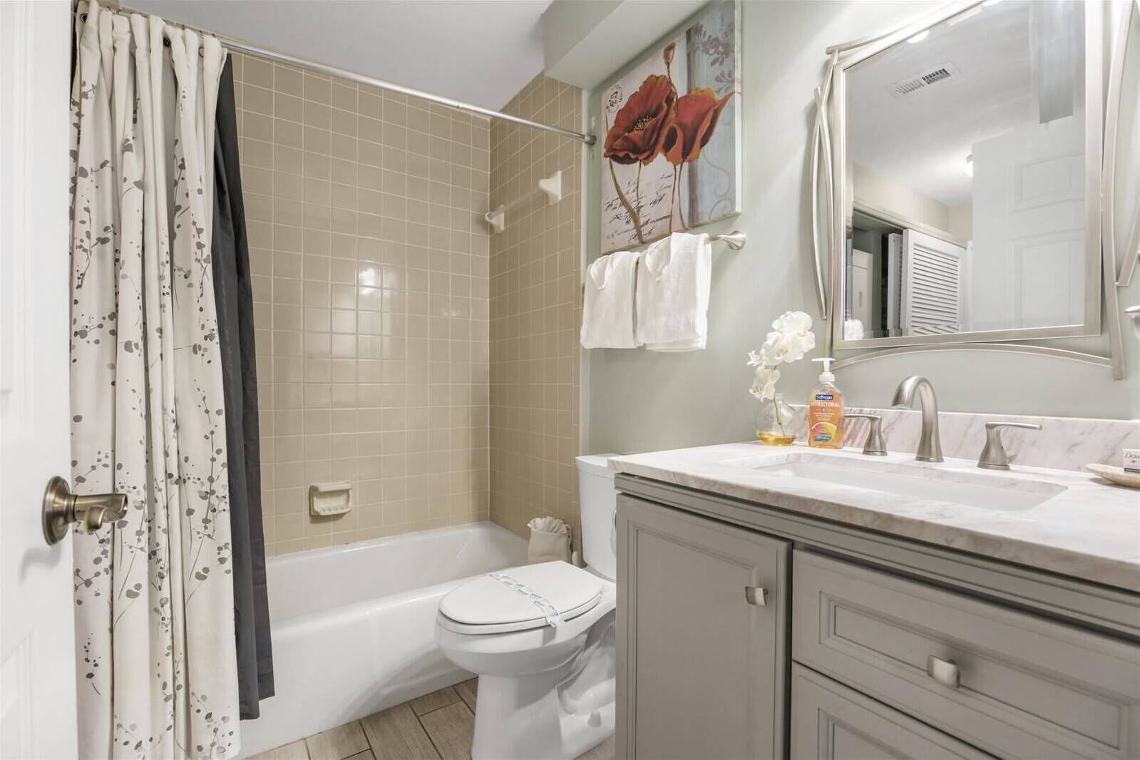 A bathroom with a bathtub, toilet, and vanity with a mirror. There are towels hanging on the wall and a flower vase on the vanity.