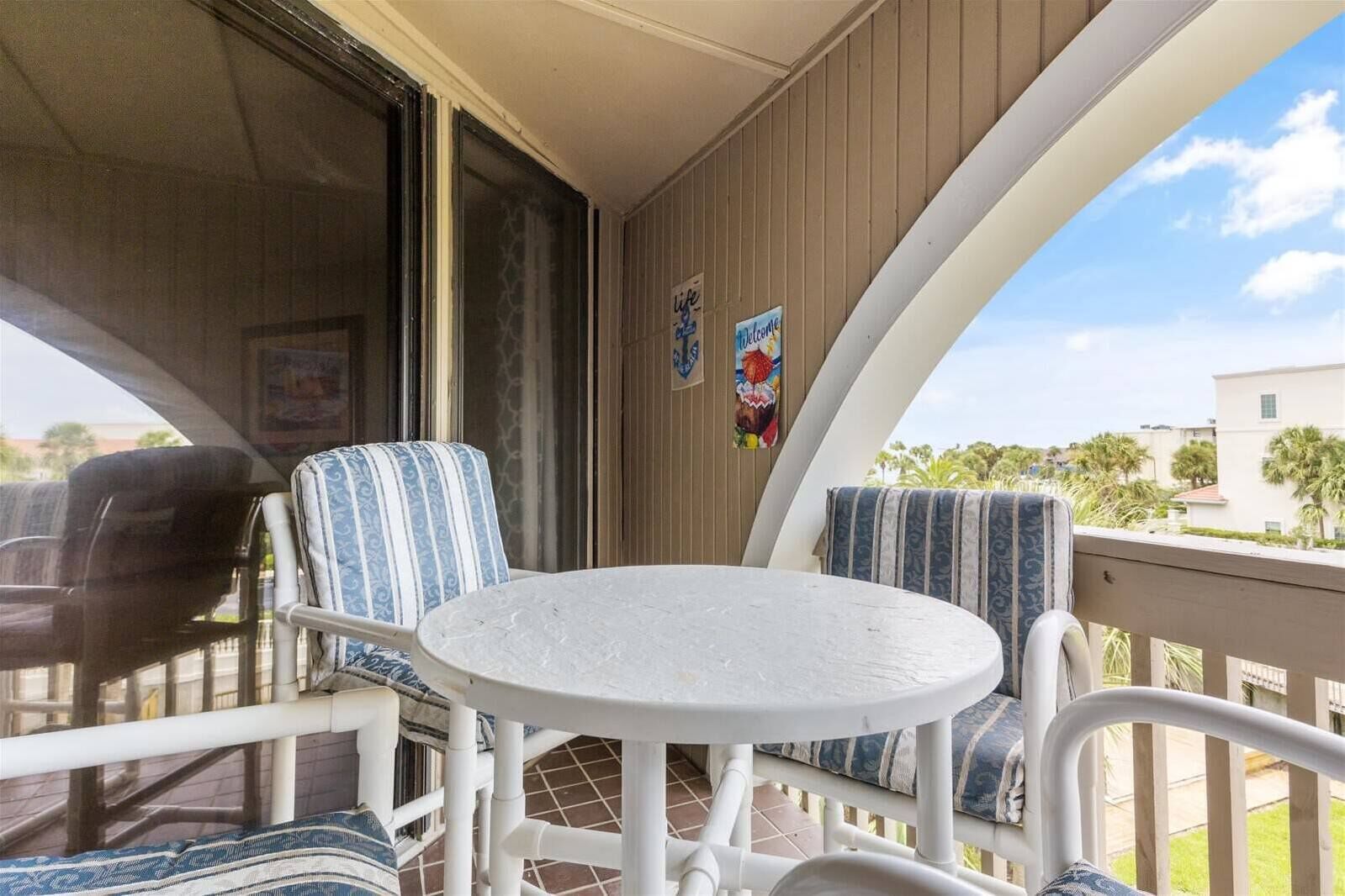 A covered outdoor seating area with a round table and two chairs, a sliding glass door, and a view of a residential area.