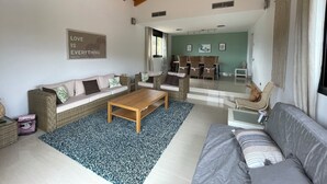 A spacious living room with a high wooden ceiling, a sofa, a coffee table, and a dining area with wicker furniture.