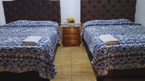A room with two beds, each with a patterned bedspread and a brown headboard. There is a wooden nightstand between the beds with a vase of yellow flowers. - Hotel San Isidro  (Pisco)