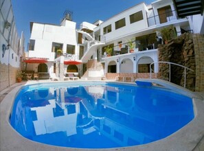 A swimming pool with a clear blue water surface, surrounded by a white building with multiple balconies and a staircase leading to an upper level. - Hotel San Isidro  (Pisco)