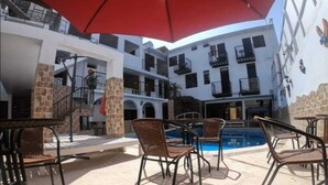 A poolside area with wicker chairs and a red umbrella, a stone pillar, and a building with multiple balconies. - Hotel San Isidro  (Pisco)