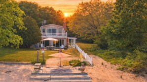 A two-story house with a deck, surrounded by trees and a sandy path leading to the entrance.