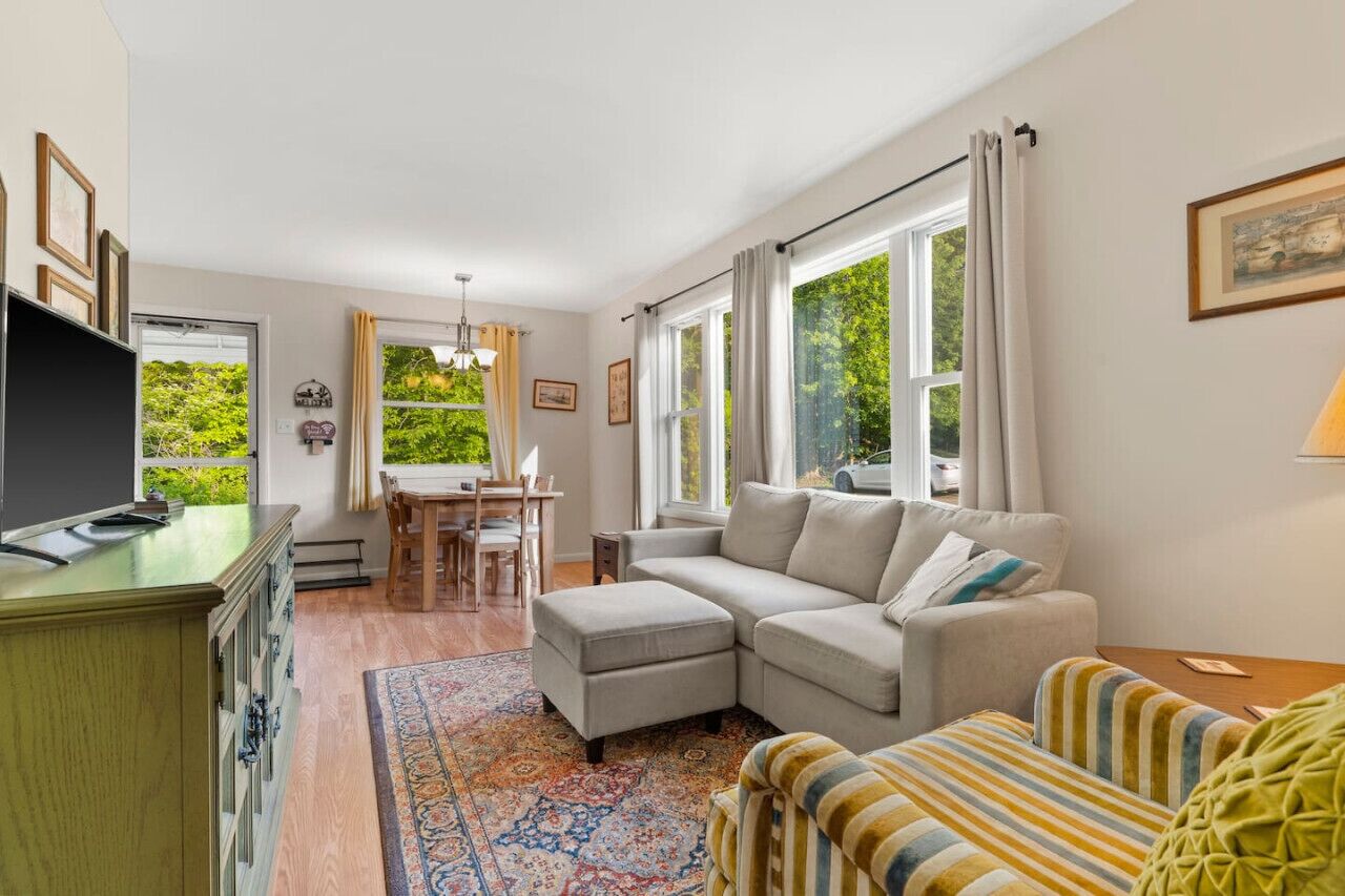 En stue med sofa, skammel og stribet lænestol, et spisebord med stole og et fjernsyn på et skab.