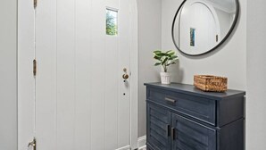 Une porte blanche avec une poignée en laiton, une commode bleu foncé dotée de tiroirs, un miroir rond, une plante en pot et un panier en osier posé sur la commode.
