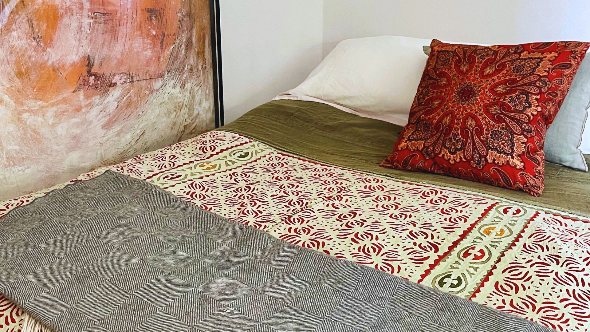Een bed met een bedsprei met patroon, een rood en goud kussen en een grijze deken.