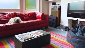 Un salon chaleureux avec un canapé rouge, un petit chien, un tapis coloré, une table basse avec des magazines, un téléviseur et un système audio.