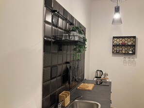 A modern kitchen with a black tiled backsplash, hanging utensils, a wooden cutting board, a kettle, and a coffee maker.
