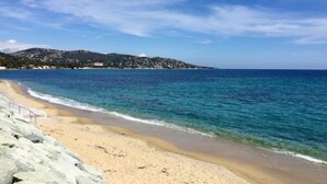 A coastal beach with clear blue waters, a sandy shore, and a hilly landscape in the background.