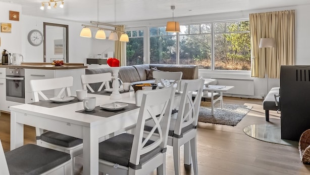 Een moderne, helder verlichte woonruimte met een eettafel, stoelen en een bank. Er is een keuken met een fornuis en een spoelbak. Grote ramen zorgen voor natuurlijk licht en uitzicht op groen buiten.