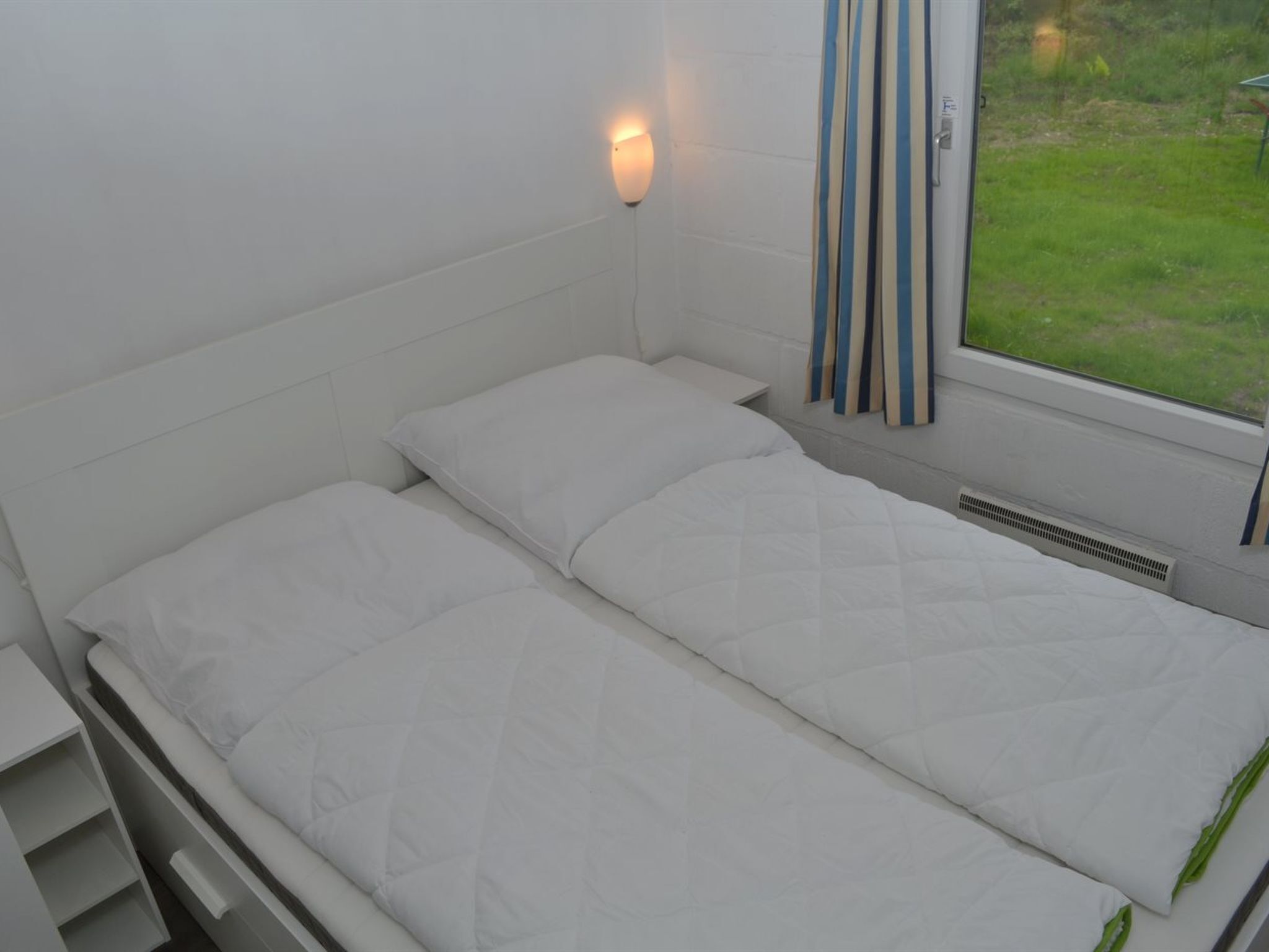 A single bed with white bedding in a room with white walls and a window with curtains.
