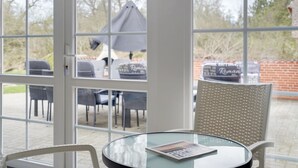 Una mesa de vidrio con una revista, un sillón de mimbre y vistas a un patio exterior con sillas y sombrillas.