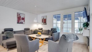 Un moderno salón con un sofá gris, una mesa de centro redonda con flores amarillas y un ventanal con vistas al exterior.