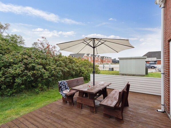 Eine Terrassengaststätte mit Holztisch und -bänken, einem großen Sonnenschirm und Blick auf Wohnhäuser im Hintergrund.