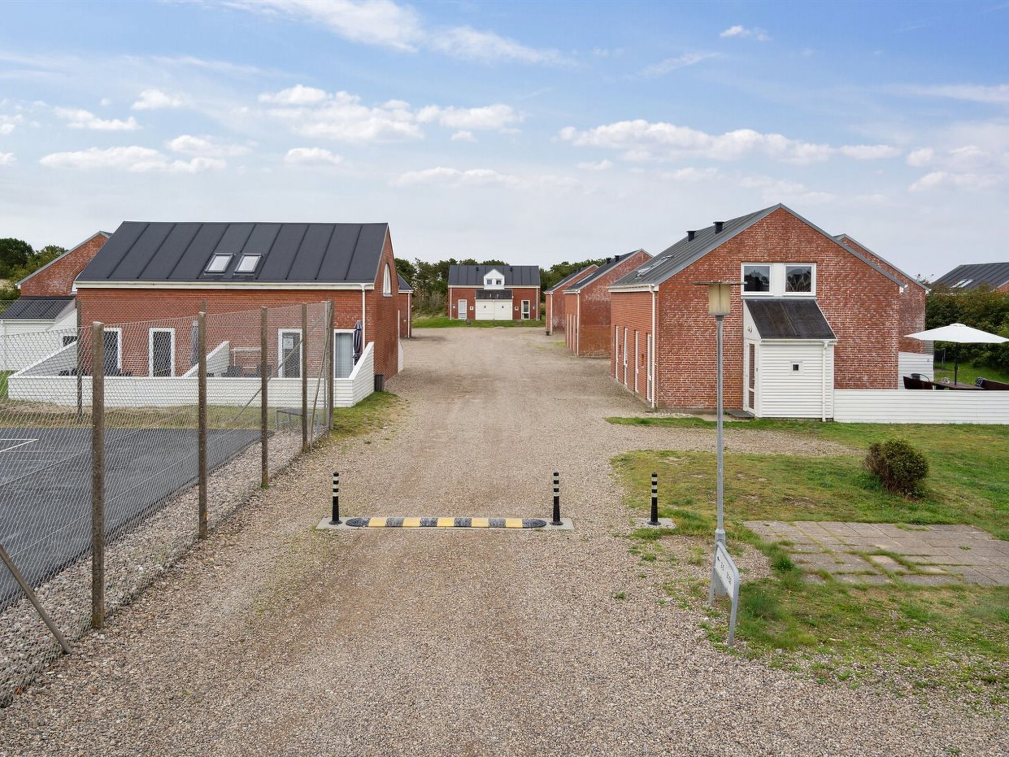 A gravel driveway leading to a row of brick buildings with a metal roof, a fenced area, and a small grassy patch with a bench.