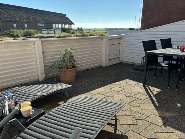 En terrasse med to liggestole, et bord med stole, en potteplante og en bygning i baggrunden.