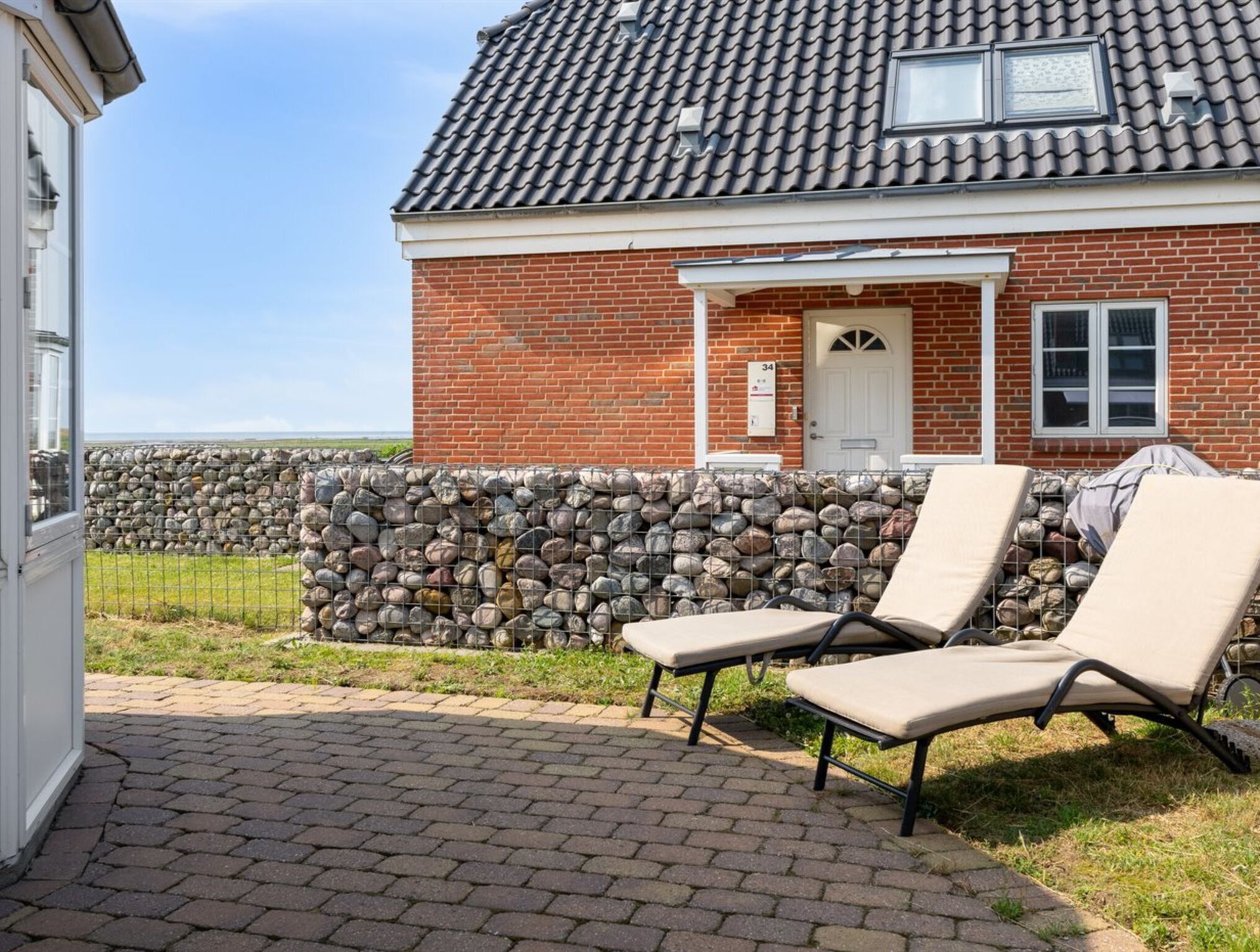 A brick house with a tiled roof, a stone wall, and a paved patio with two lounge chairs.
